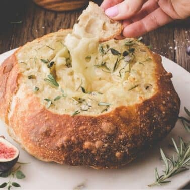 A hand dips bread into a round loaf hollowed out and filled with melted cheese, garnished with herbs, with figs and rosemary nearby on a marble platter.