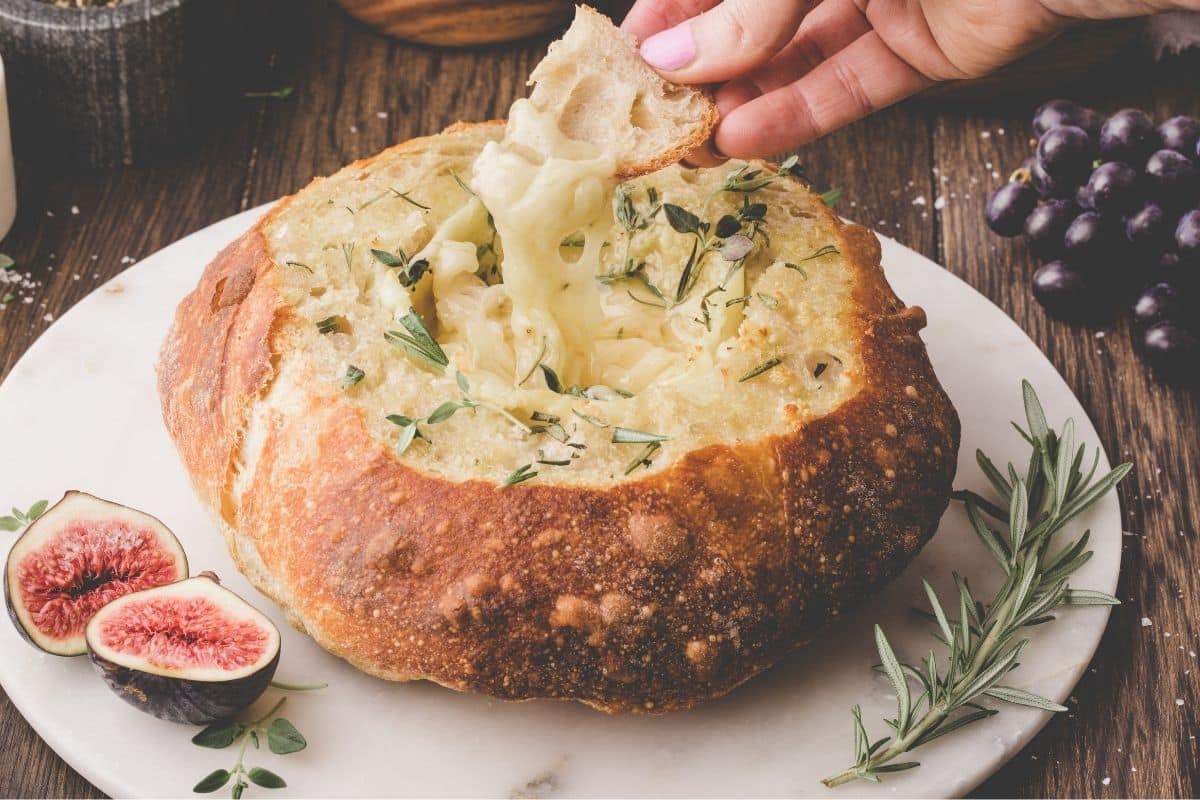 A hand dips bread into a round loaf hollowed out and filled with melted cheese, garnished with herbs, with figs and rosemary nearby on a marble platter.