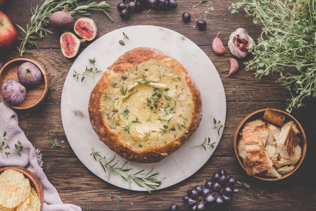 A round loaf of bread filled with baked cheese sits on a marble board, surrounded by figs, grapes, herbs, apple, garlic, chips, and a bowl of bread pieces.