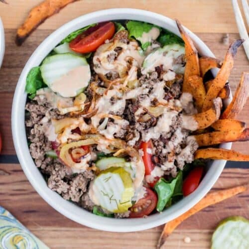 A bowl filled with ground beef, lettuce, tomato, sliced cucumber, pickles, crispy onions, dressing, and sweet potato fries on the side, set on a wooden surface.