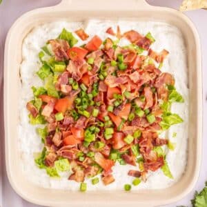 A square dish filled with a creamy dip, topped with chopped lettuce, tomatoes, bacon, and green onions.