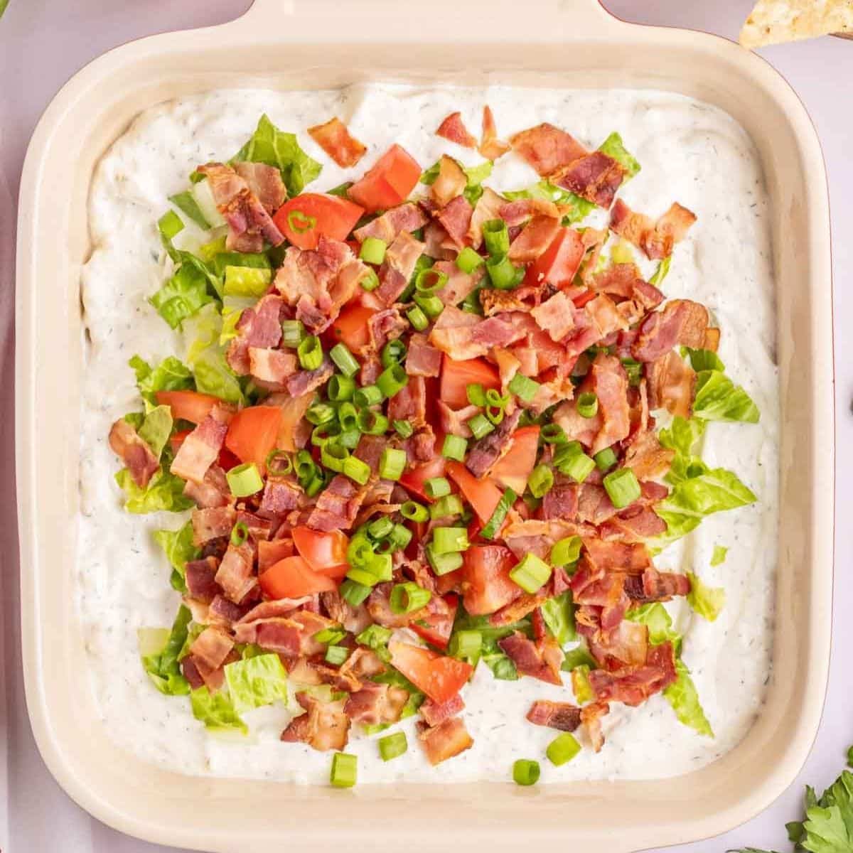 A square dish filled with a creamy dip, topped with chopped lettuce, tomatoes, bacon, and green onions.