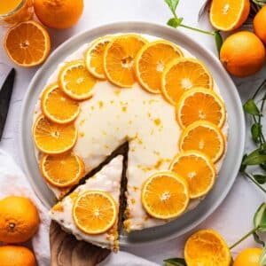 A round iced orange clementine cake on a white plate, decorated with orange slices on top. One slice is being removed. Whole and halved oranges are around the plate.