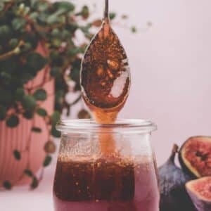 A spoon drizzling fig jam into a glass jar, with fresh figs and a potted plant in the background.