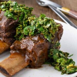 Braised beef short ribs served on a white plate, topped with a green herb sauce, with utensils in the background.