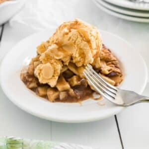A slice of apple pie topped with a scoop of vanilla ice cream and caramel sauce on a white plate, with a fork resting on the dessert.