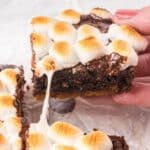 A hand holds a s'mores brownie topped with toasted marshmallows and melted chocolate, showing a gooey marshmallow stretch.