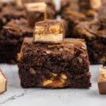 A close-up of a snickers brownie with chopped nuts, topped with a piece of caramel and nougat candy, with more brownies in the background.