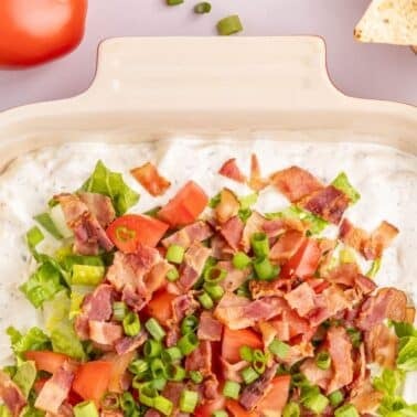A baking dish filled with a creamy dip topped with chopped lettuce, tomatoes, bacon, and green onions; tortilla chips and a tomato are nearby.