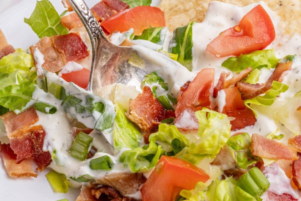 A close-up of a salad with lettuce, tomatoes, bacon, green onions, and creamy dressing, with a fork partially visible.