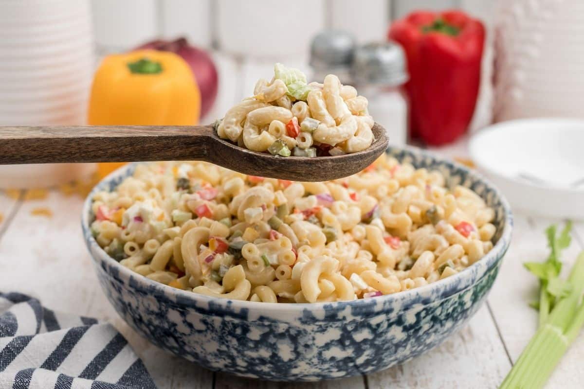 A wooden spoon holds a serving of macaroni salad above a bowl filled with the same salad, surrounded by fresh vegetables and kitchen items.