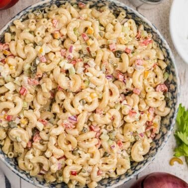 A large bowl of macaroni salad with chopped vegetables, including red bell pepper and celery, on a wooden table surrounded by ingredients and utensils.