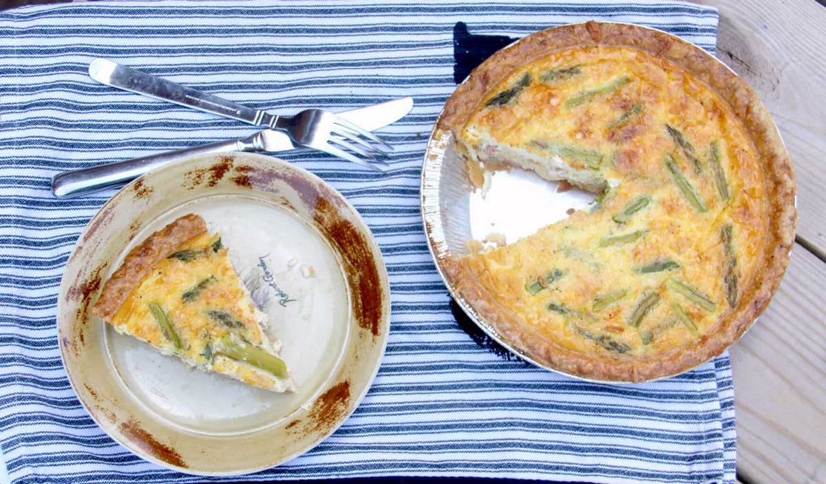 A quiche with a slice removed sits on a striped cloth; the slice is served on a plate with a fork and knife beside it.