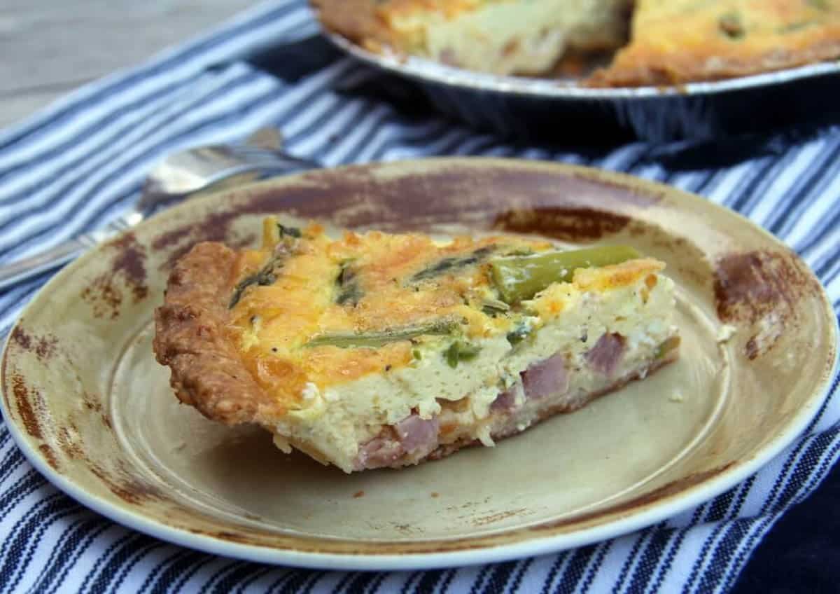 A slice of quiche with ham, cheese, and green vegetables on a rustic beige plate, with a striped cloth and utensils in the background.
