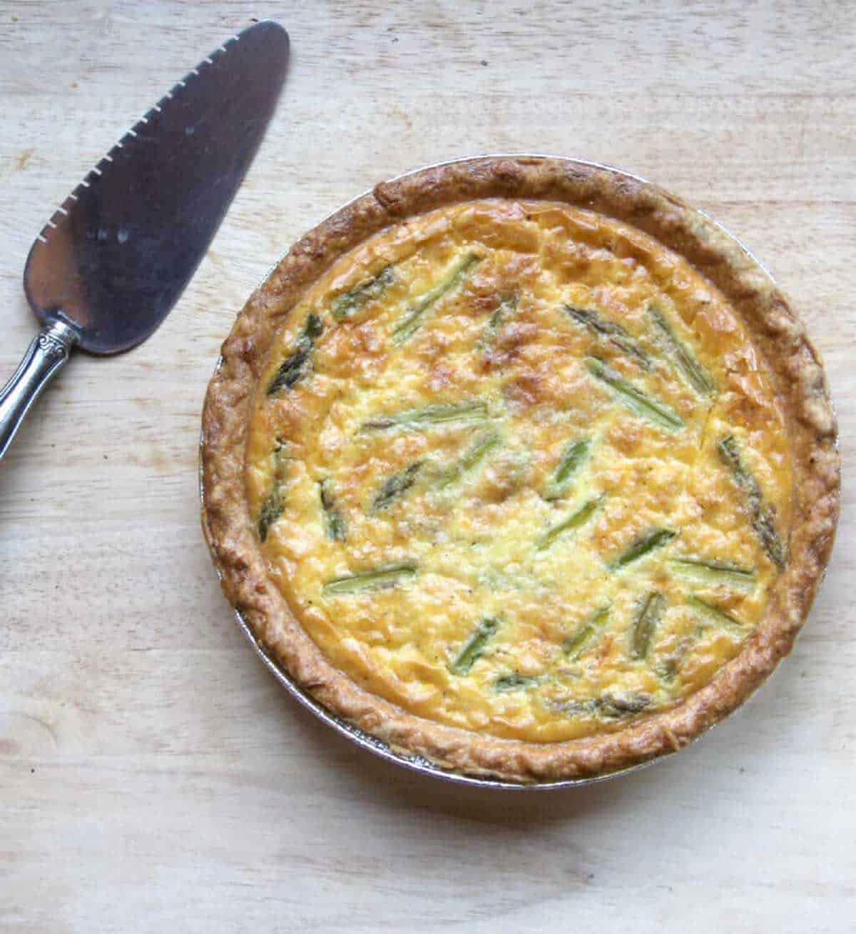 A bacon cheddar quiche with a golden crust and visible asparagus pieces sits on a wooden surface next to a metal pie server.