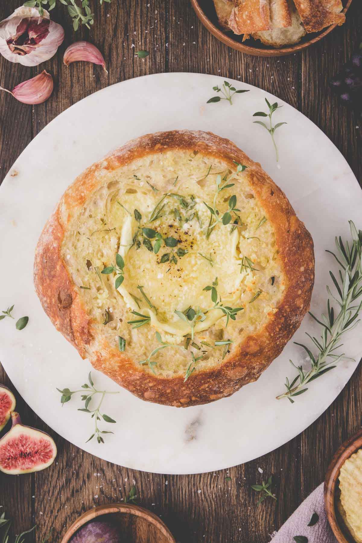 A round loaf of bread with baked brie cheese in the center, garnished with fresh herbs, placed on a white marble board with figs, garlic, and bread slices nearby.