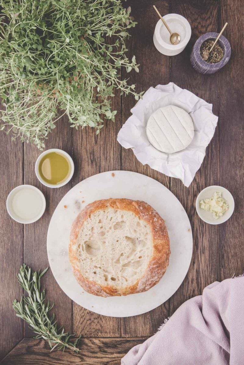 A round loaf of bread on a marble board with a wheel of cheese, small bowls of oil, garlic, salt, and pepper, fresh herbs, and a lilac cloth on a wooden table.