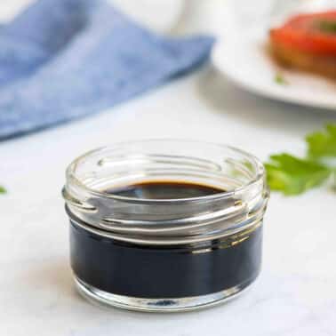 A small glass jar filled with balsamic glaze, sits on a white surface with herbs and a blurred plate in the background.