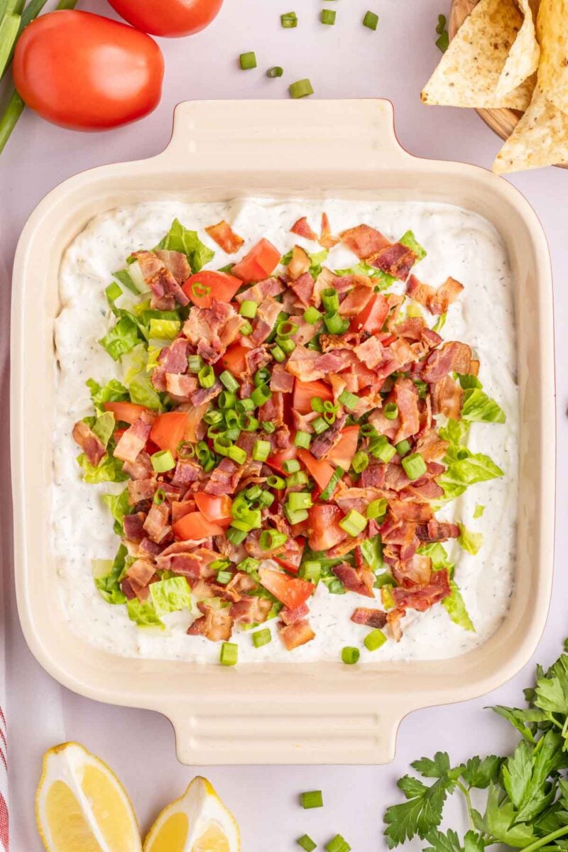A square dish filled with BLT dip topped with chopped lettuce, tomatoes, bacon, and green onions, surrounded by fresh vegetables, herbs, lemon wedges, and tortilla chips.