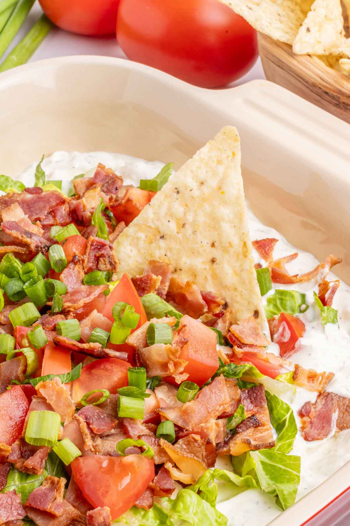 A close-up of a BLT dip topped with chopped bacon, tomatoes, and green onions, with a tortilla chip inserted into the dip. Fresh tomatoes and chips are visible in the background.