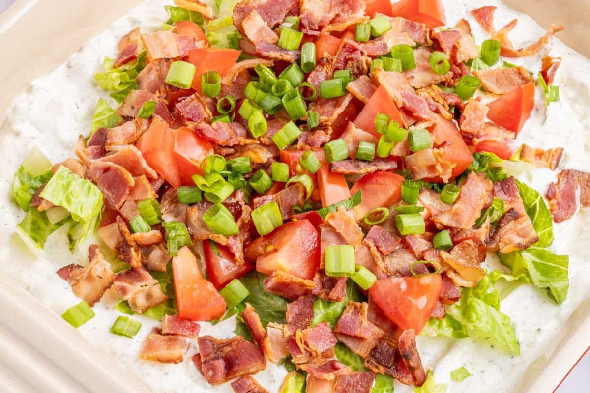 Chopped bacon, tomatoes, and green onions on a creamy layer over chopped lettuce in a dish.
