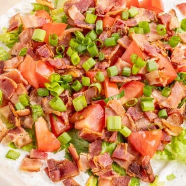 A close-up of a layered salad topped with chopped bacon, diced tomatoes, and sliced green onions over a creamy base.