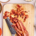Sliced and chopped cooked bacon on a wooden cutting board with a knife, surrounded by bowls of salt, mayonnaise, and pepper.