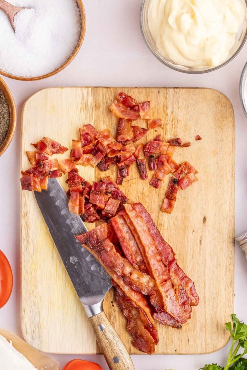 Sliced and chopped cooked bacon on a wooden cutting board with a knife, surrounded by bowls of salt, mayonnaise, and pepper.
