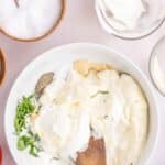 A bowl with mayonnaise, sour cream, herbs, and seasonings being mixed with a wooden spoon, surrounded by salt, a glass with sour cream, and fresh ingredients.