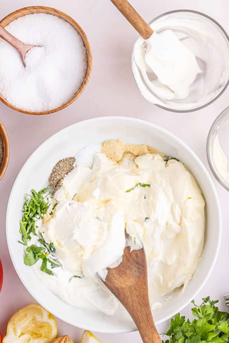 A bowl with mayonnaise, sour cream, herbs, and seasonings being mixed with a wooden spoon, surrounded by salt, a glass with sour cream, and fresh ingredients.