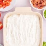 A baking dish filled with a creamy white mixture, surrounded by bowls of chopped tomatoes, crumbled bacon, chopped green onions, and fresh herbs.