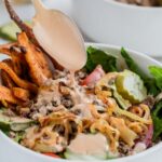 A spoon drizzles creamy dressing over a bowl of salad with ground meat, sweet potato fries, cucumbers, greens, and grilled onions.