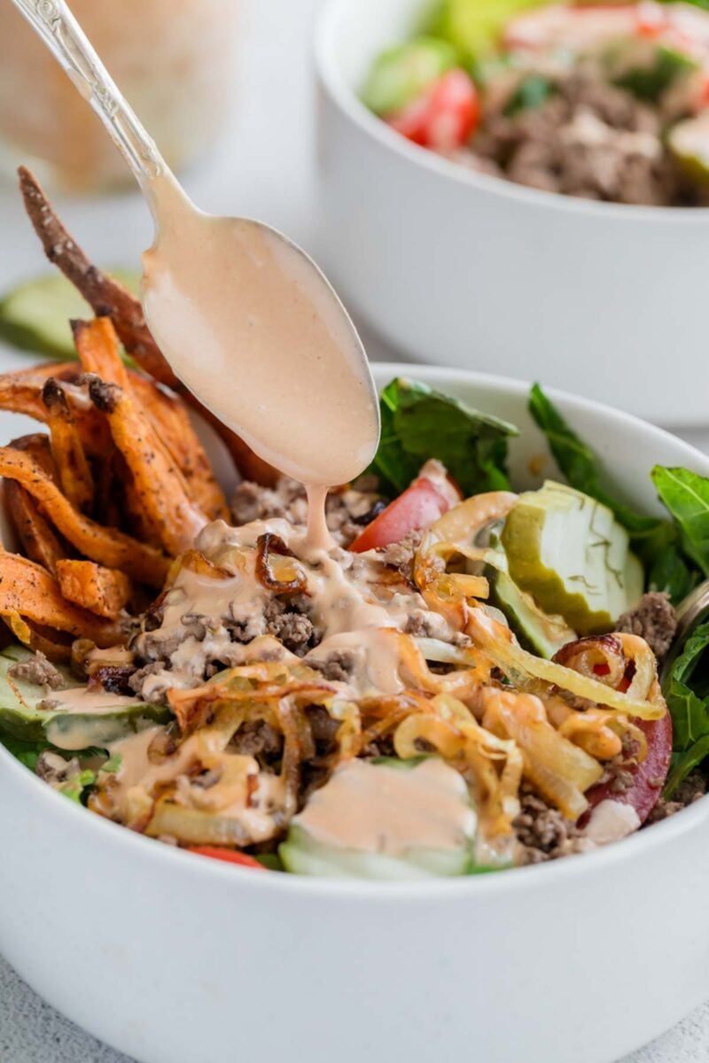 A spoon drizzles creamy dressing over a bowl of salad with ground meat, sweet potato fries, cucumbers, greens, and grilled onions.