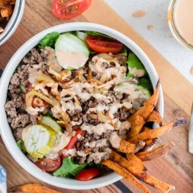 A burger bowl with ground beef, lettuce, tomato, pickles, caramelized onions, and sauce, topped with sweet potato fries; a fork, napkin, and extra fries are nearby.