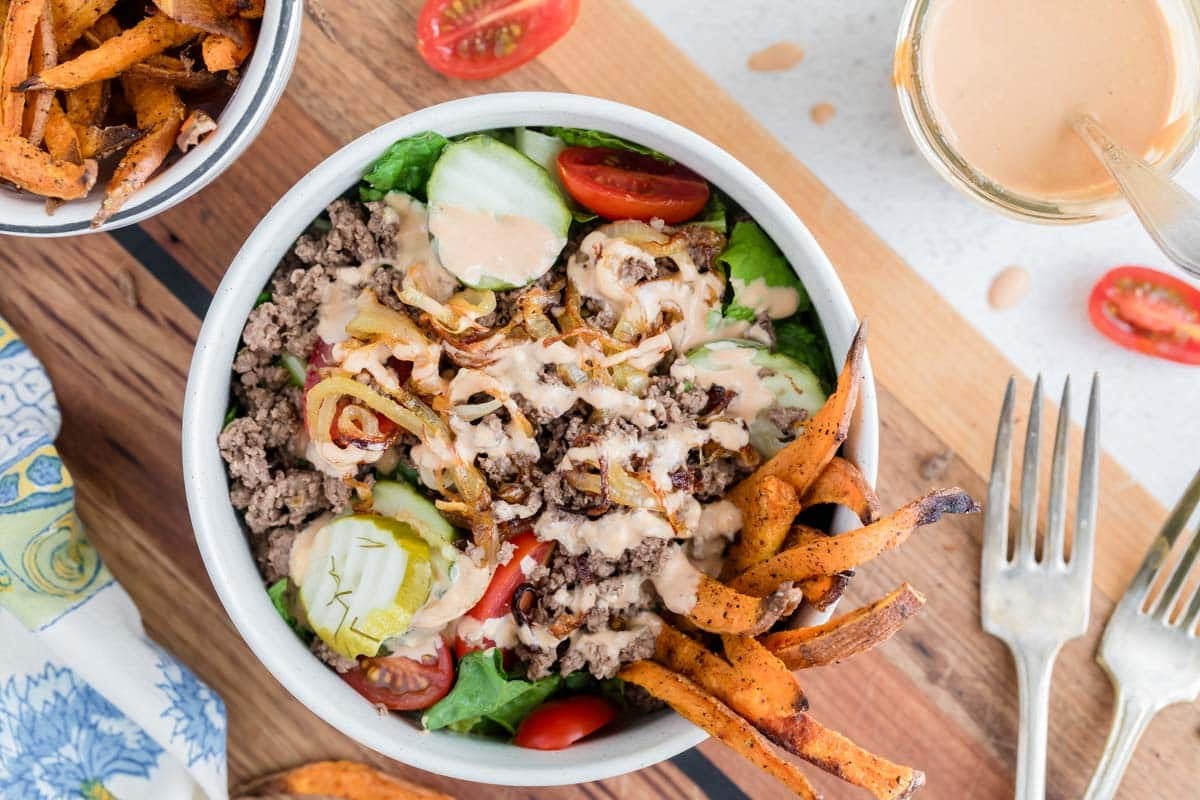 A burger bowl with ground beef, lettuce, tomato, pickles, caramelized onions, and sauce, topped with sweet potato fries; a fork, napkin, and extra fries are nearby.