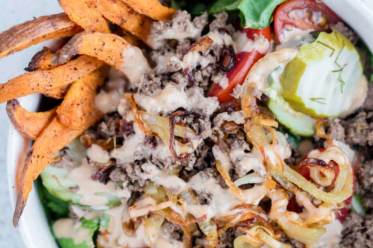A bowl containing ground beef, sliced pickles, tomatoes, lettuce, grilled onions, sweet potato fries, and a drizzle of creamy sauce.