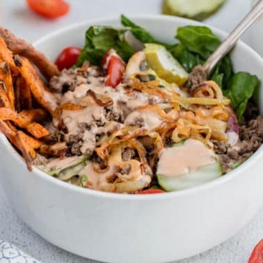 A white bowl filled with ground beef, sliced cucumbers, cherry tomatoes, pickles, greens, sweet potato fries, sautéed onions, and a drizzle of creamy sauce.