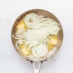 A stainless steel pan with sliced onions and chunks of butter beginning to cook on a white background.