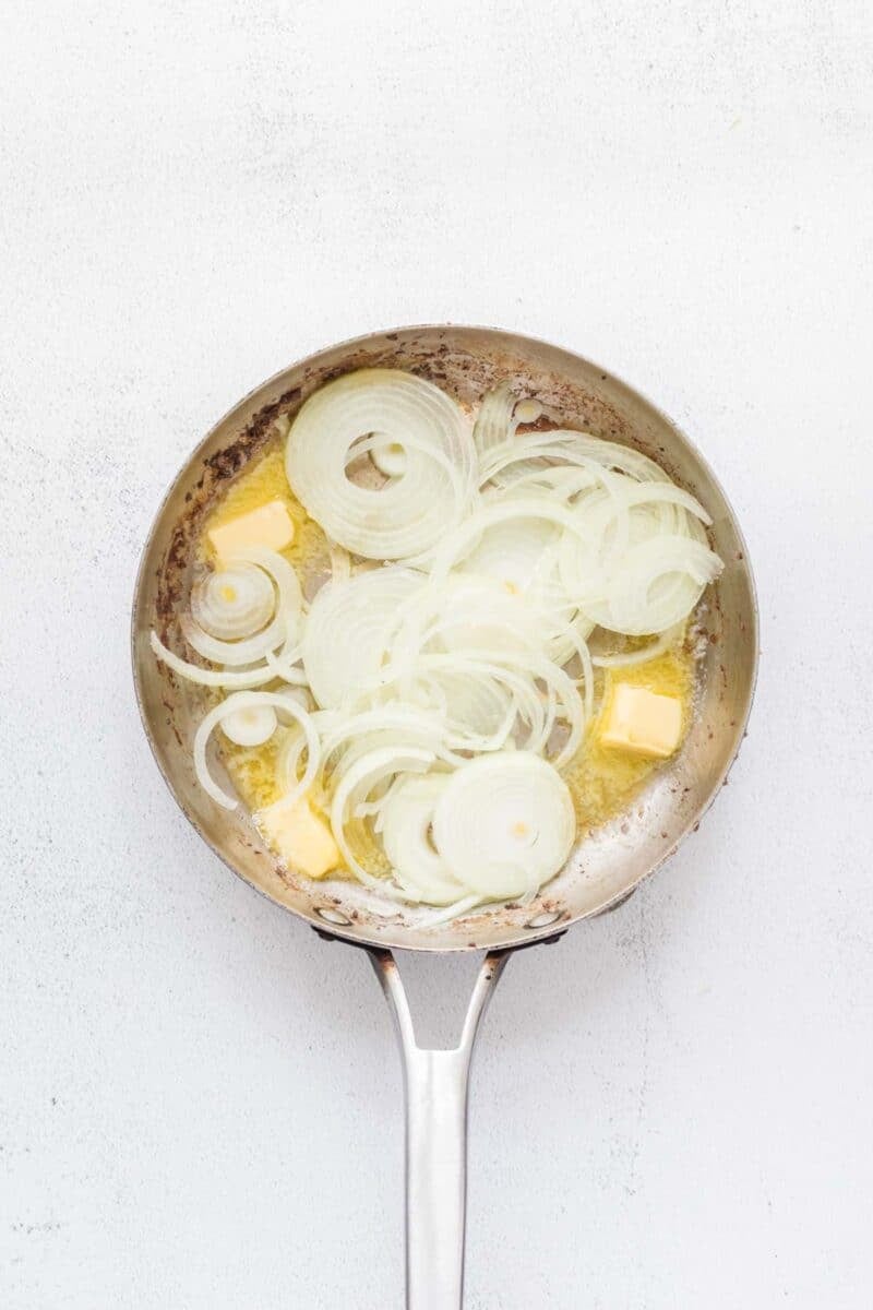 A stainless steel pan with sliced onions and chunks of butter beginning to cook on a white background.
