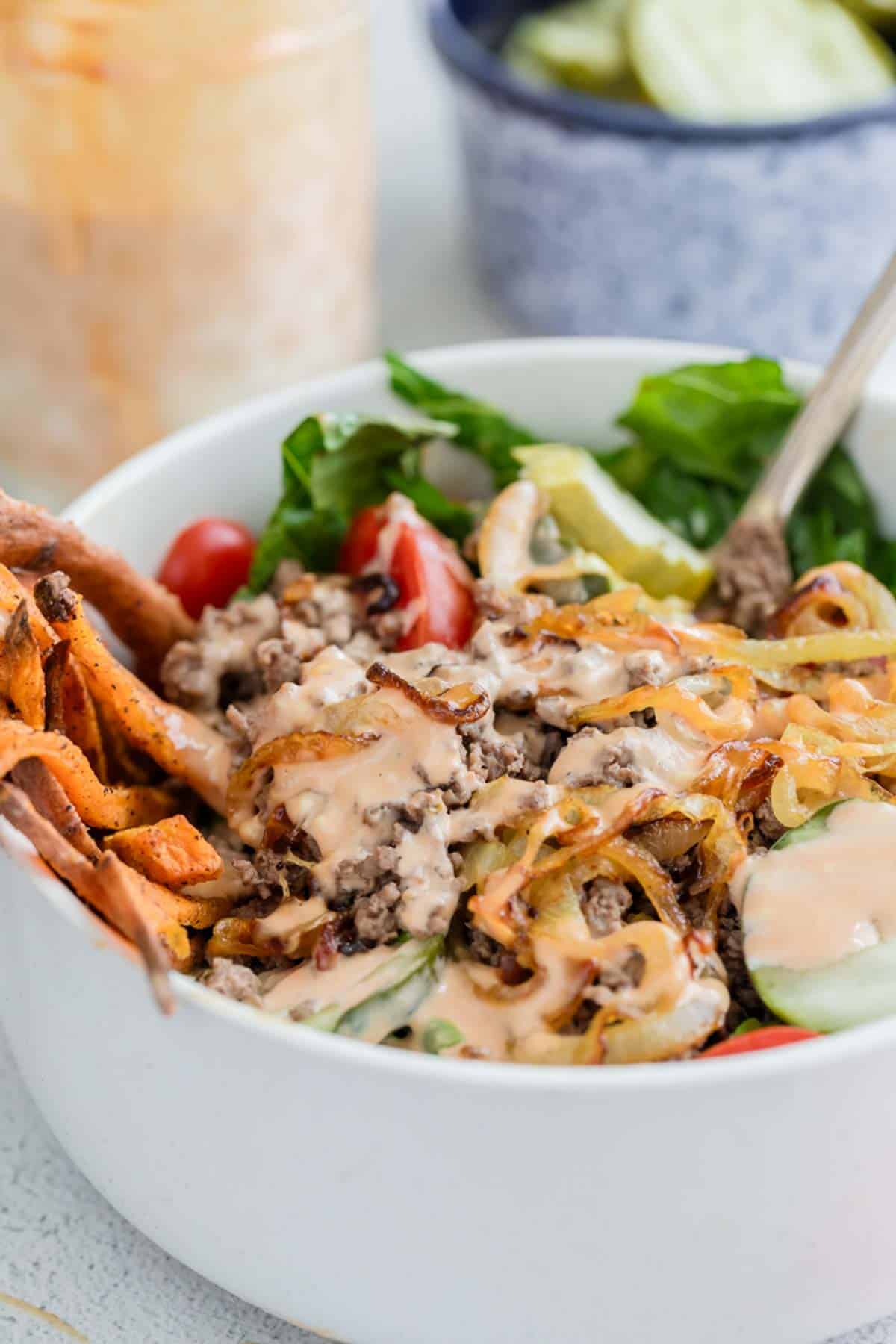 A white bowl filled with ground beef, lettuce, tomatoes, pickles, sweet potato fries, and drizzled sauce, with a fork inside.
