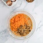 A glass bowl with cake batter, shredded carrots, and chopped walnuts sits on a marble countertop. Next to it are cracked egg shells in a dish and a small bowl of walnuts.