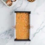 A loaf pan filled with carrot cake batter sits on a marble surface, with cracked eggshells and a bowl of walnuts nearby.