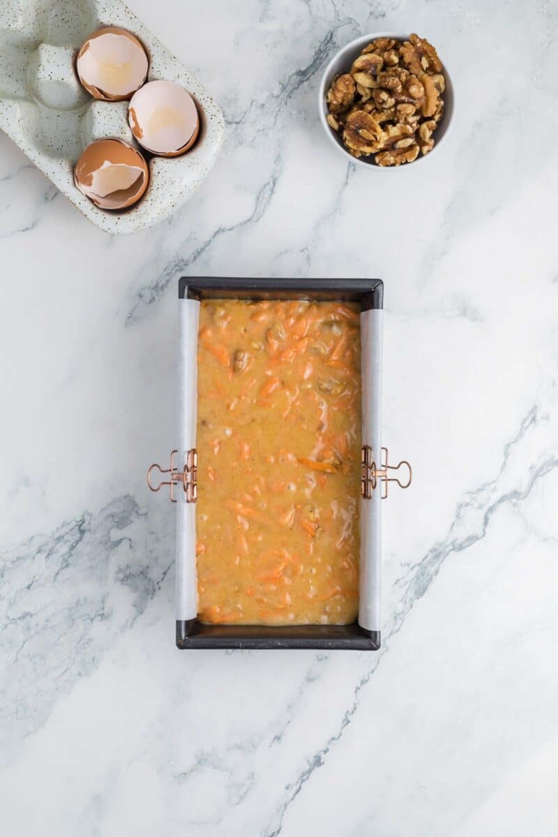 A loaf pan filled with carrot cake batter sits on a marble surface, with cracked eggshells and a bowl of walnuts nearby.