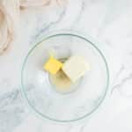 A glass bowl with a block of cream cheese and a chunk of butter on a marble surface, next to a bowl of walnuts and a beige cloth.
