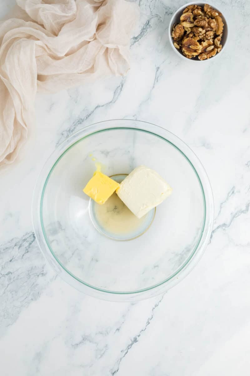A glass bowl with a block of cream cheese and a chunk of butter on a marble surface, next to a bowl of walnuts and a beige cloth.