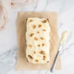 A loaf cake with white frosting and walnut halves on top sits on parchment paper, beside a bowl of walnuts and a knife with frosting on a marble surface.