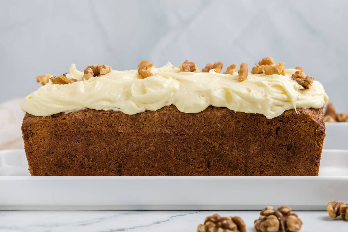 A loaf of carrot cake topped with cream cheese frosting and walnut halves sits on a white rectangular plate.