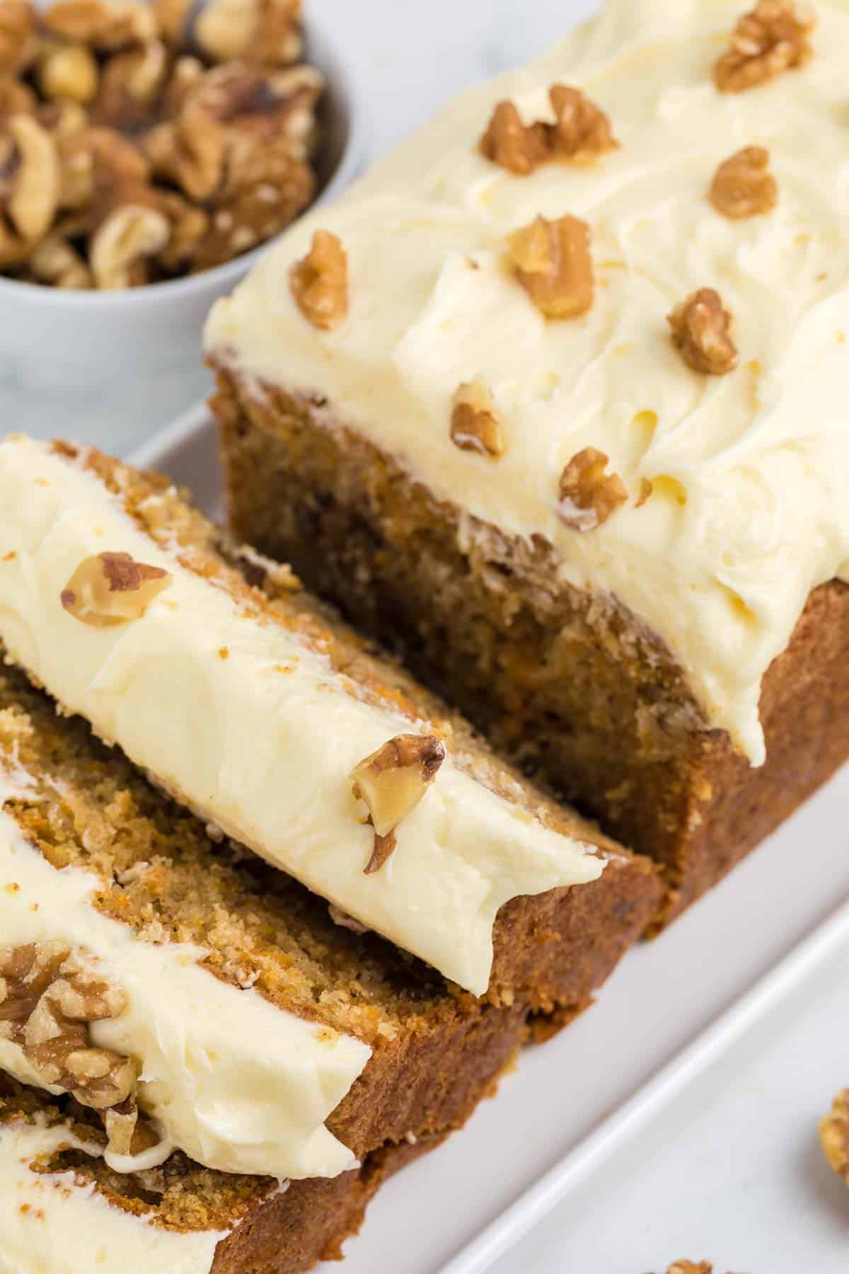 Sliced loaf of carrot cake topped with cream cheese frosting and walnut pieces, with additional walnuts in a bowl in the background.