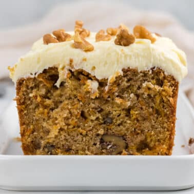 A sliced loaf of carrot cake with cream cheese frosting and walnut pieces on top, displayed on a white rectangular plate.