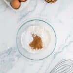 A glass bowl with flour, baking powder, and cinnamon on a marble surface, with eggs, walnuts, and a metal whisk nearby.